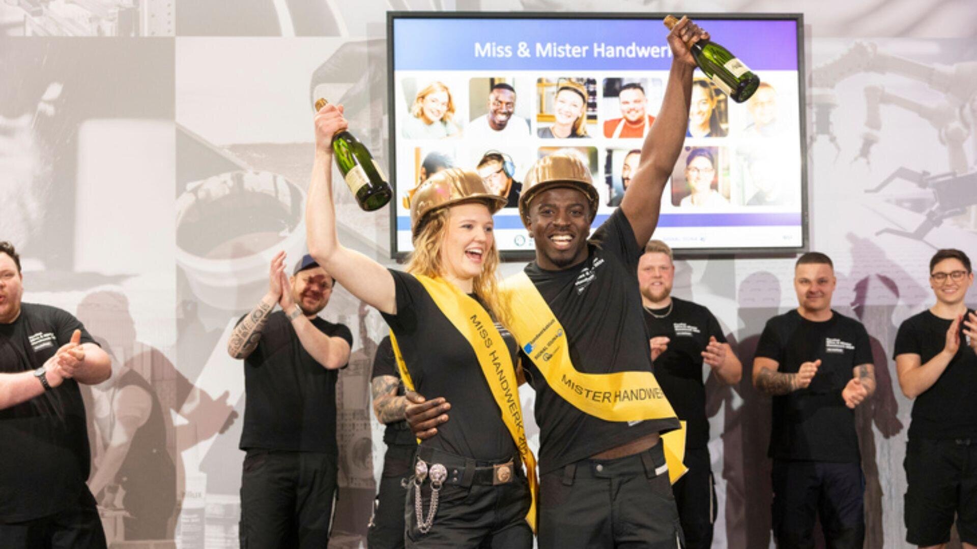 Zwei lächelnde Gewinner mit goldenen Bauhelmen und Schärpen halten Champagnerflaschen hoch und feiern auf der Bühne, während die Leute hinter ihnen klatschen. Im Hintergrund ist auf einem Bildschirm „Miss & Mister Handwerk“ zu sehen.