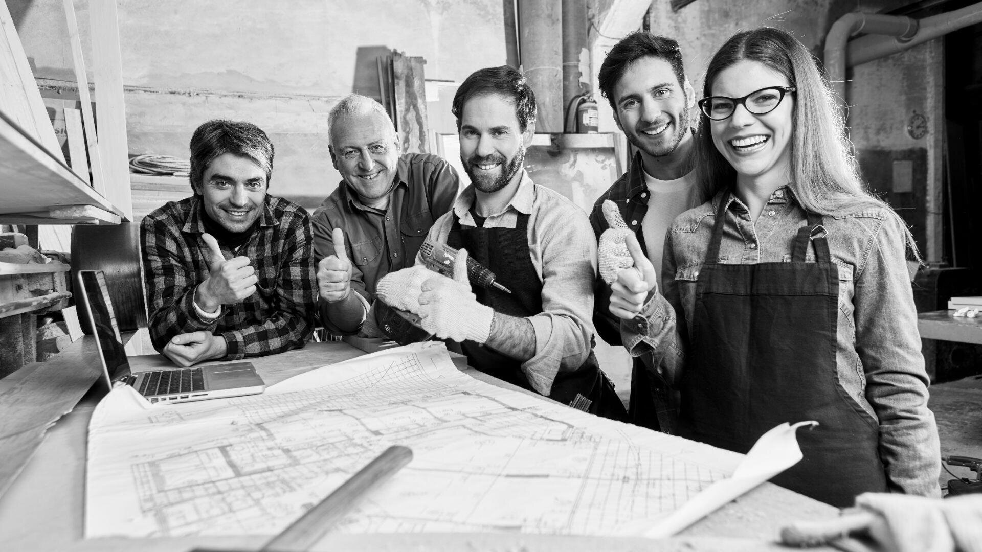 Fünf Personen in Schürzen und Freizeitkleidung stehen lächelnd und mit erhobenem Daumen um einen Arbeitstisch mit Bauplänen in einer Werkstattumgebung zusammen.