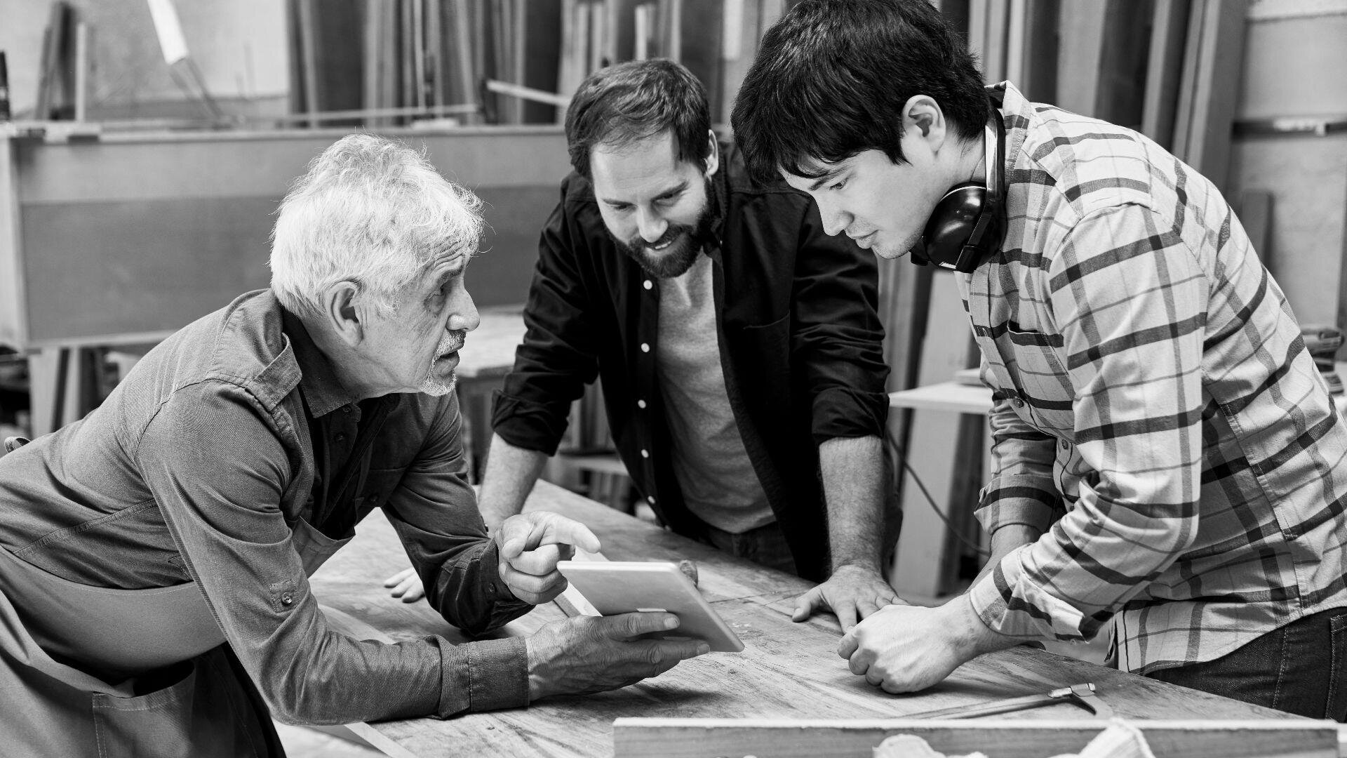 Drei Männer in einer Holzwerkstatt schauen gemeinsam auf ein Tablet auf einer Werkbank. Ein älterer Mann zeigt auf den Bildschirm, während zwei jüngere Männer, einer davon mit Kopfhörern um den Hals, aufmerksam zuschauen.