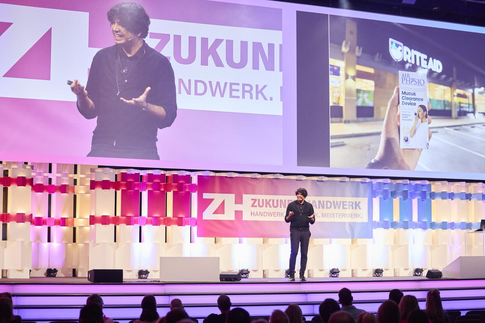 Ein Redner steht auf einer hell erleuchteten Bühne vor einem Publikum, hinter ihm sind auf großen Bildschirmen das Logo von Zukunft Handwerk und das Bild einer Hand zu sehen, die ein Produkt von Rite Aid in einer Apotheke hält.