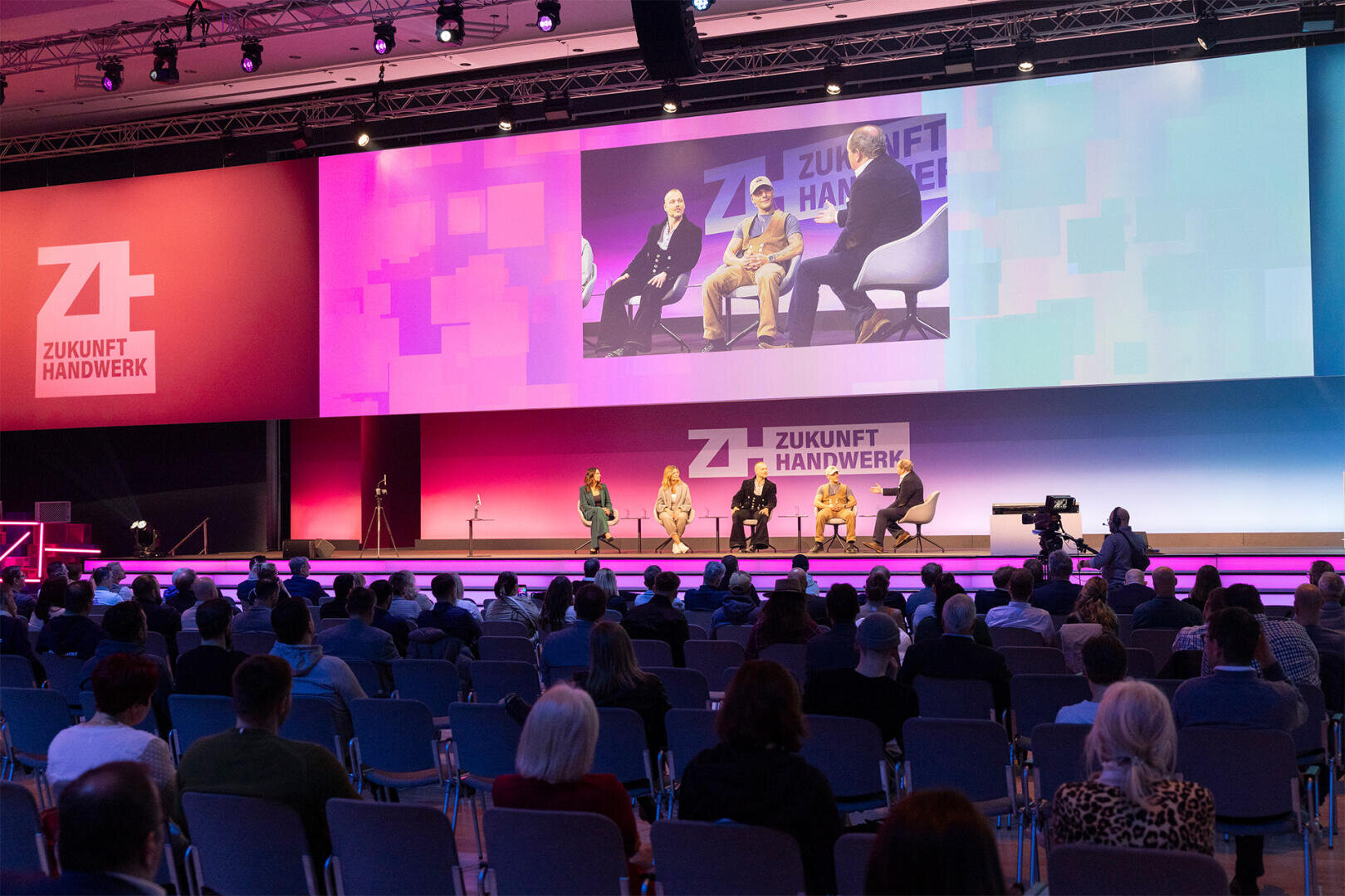 Eine fünfköpfige Diskussionsrunde sitzt auf der Bühne der Veranstaltung Zukunft Handwerk, vor ihr sitzt das Publikum und über der Bühne ist eine große Leinwand mit den Diskussionsteilnehmern zu sehen.