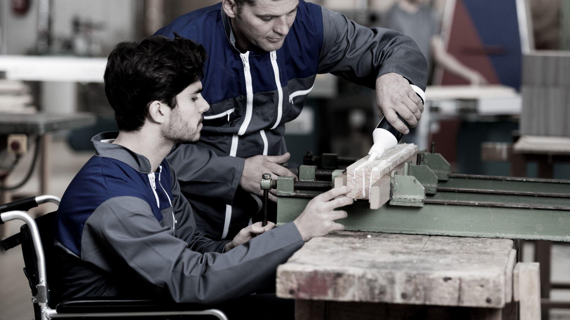 Ein Mann im Rollstuhl und ein anderer Mann arbeiten gemeinsam an einer Werkbank und spannen in einer Werkstatt ein Stück Holz in einem Schraubstock ein. Beide tragen Arbeitskleidung.