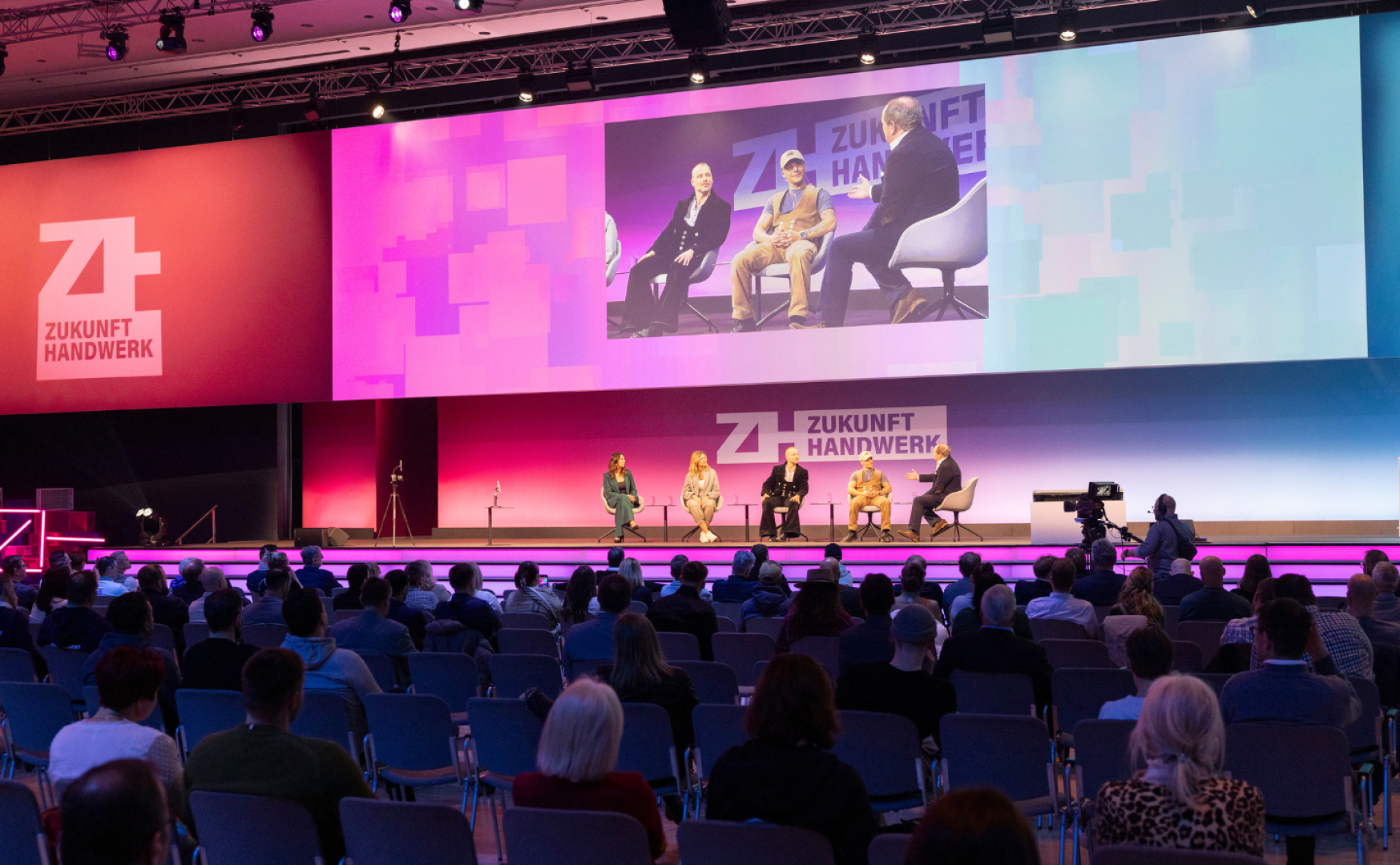 Eine fünfköpfige Gruppe spricht auf der Bühne einer Konferenz mit dem Branding Zukunft Handwerk. Das Publikum sitzt mit Blick auf die Bühne, und ein Kameramann zeichnet die Veranstaltung auf. Die Bühne ist mit lila und blauen Lichtern beleuchtet.