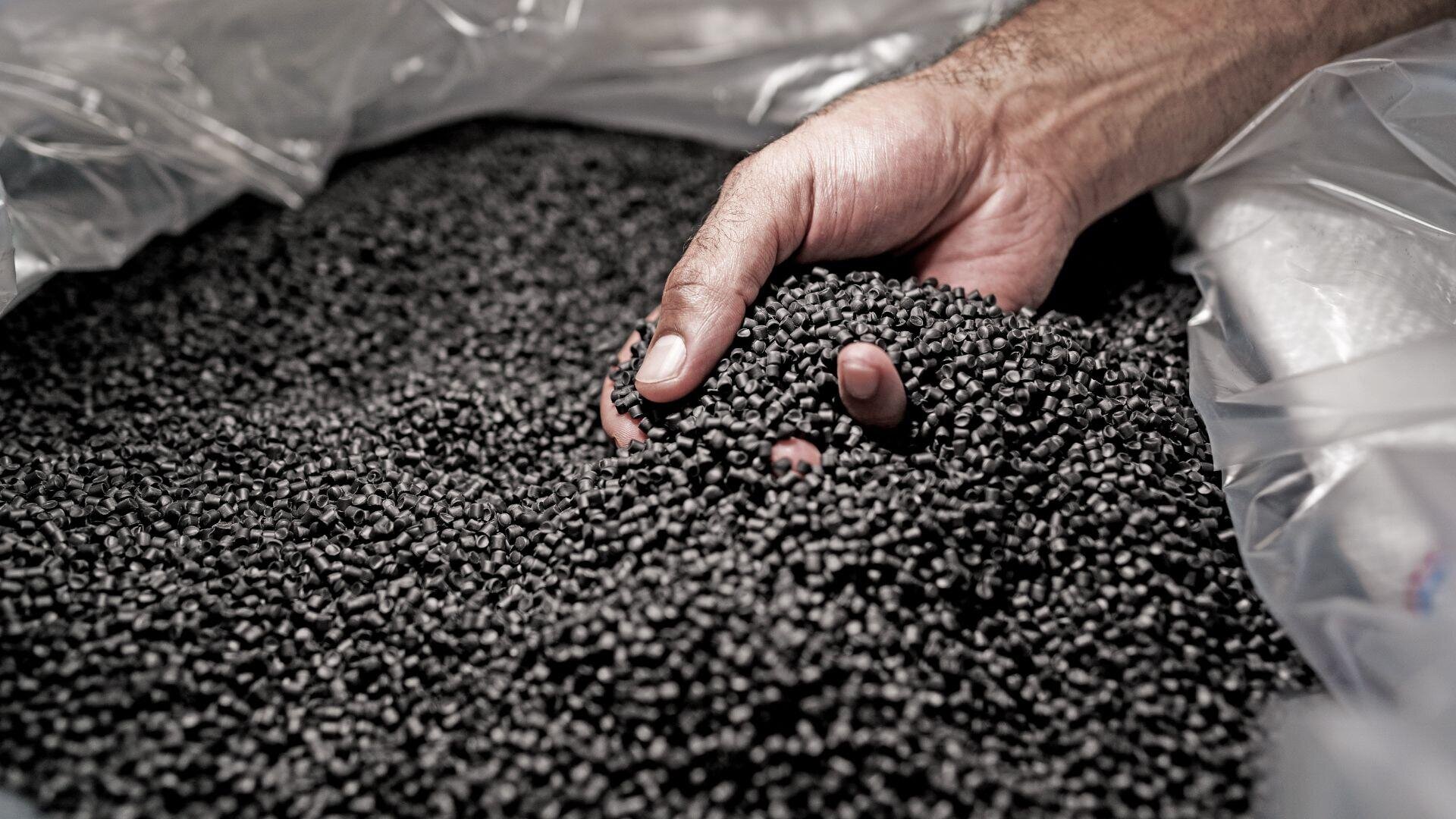 Eine Nahaufnahme einer Hand, die schwarze Plastikpellets aus einer großen durchsichtigen Plastiktüte schöpft, wobei die Pellets dicht um die Hand gepackt sind.
