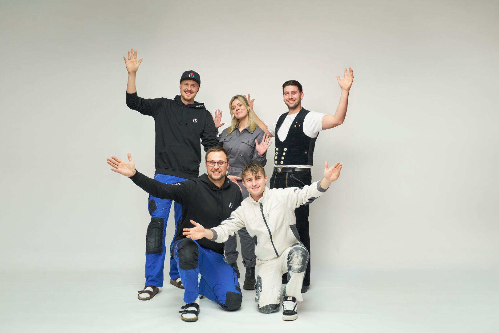 Fünf Personen posieren gemeinsam vor einem weißen Hintergrund, lächeln und heben ihre Hände in die Luft. Zwei knien vor ihnen, während drei hinter ihnen stehen. Sie tragen legere Kleidung und Arbeitskleidung.