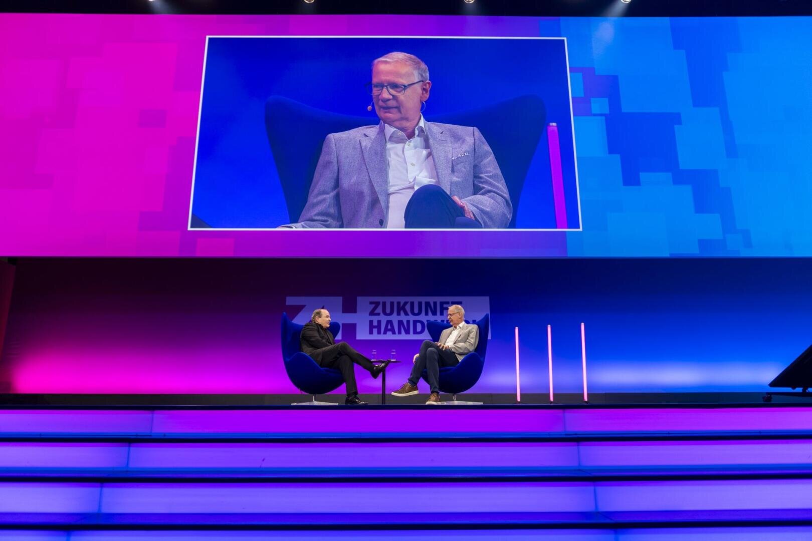 Zwei Personen sitzen auf Stühlen auf der Bühne und diskutieren. Über ihnen ist ein Mann auf einer großen Leinwand zu sehen. Der Bühnenhintergrund ist blau und rosa, und hinter ihnen steht ein Schild mit der Aufschrift „Zukunft Hand“.
