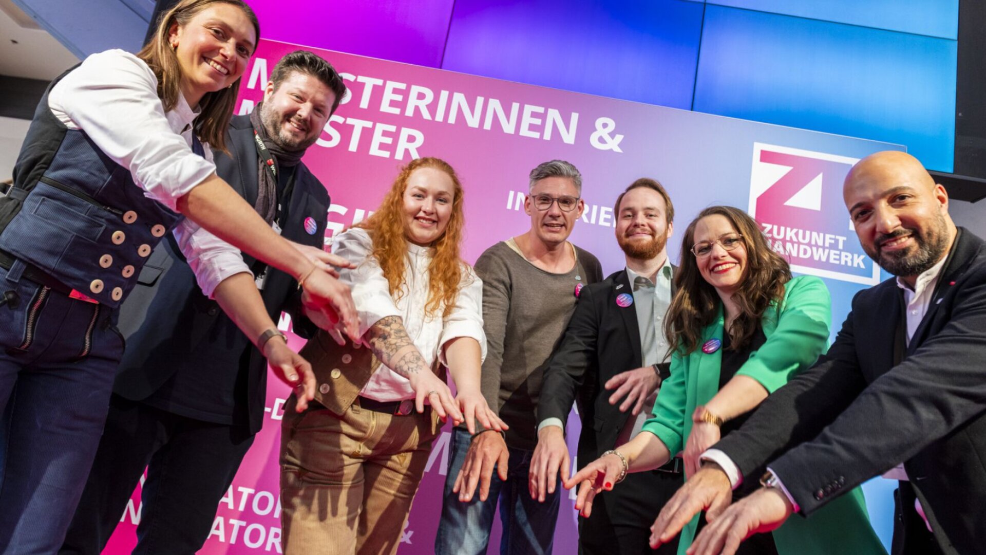 Eine Gruppe von sieben lächelnden Menschen steht mit ausgestreckten Händen im Halbkreis vor der farbenfrohen Kulisse der Veranstaltung „Zukunft Handwerk“. Sie wirken begeistert und unterstützend.