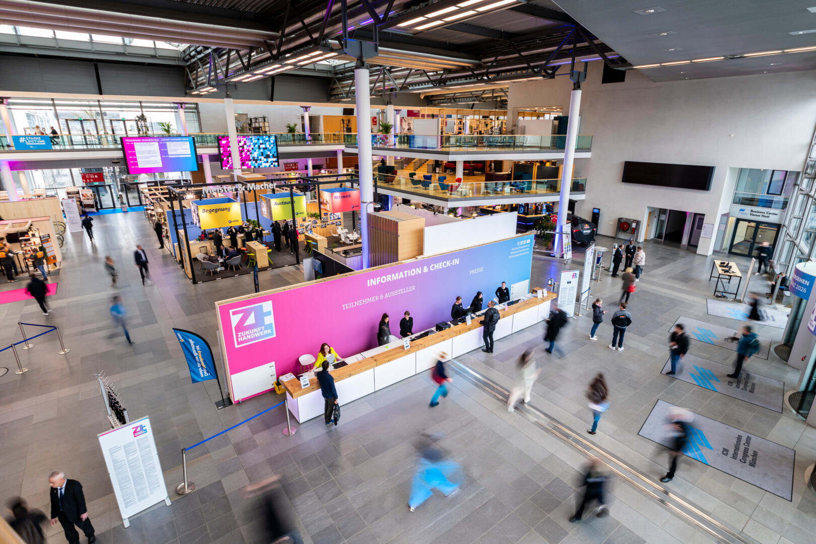 Ein geschäftiges, modernes Foyer eines Kongresszentrums mit einem Informations- und Check-in-Schalter, bunten Ständen, Menschen, die herumlaufen, und mehreren Stockwerken. Schilder, Bildschirme und offene Räume deuten auf eine organisierte Veranstaltung oder Konferenz hin.