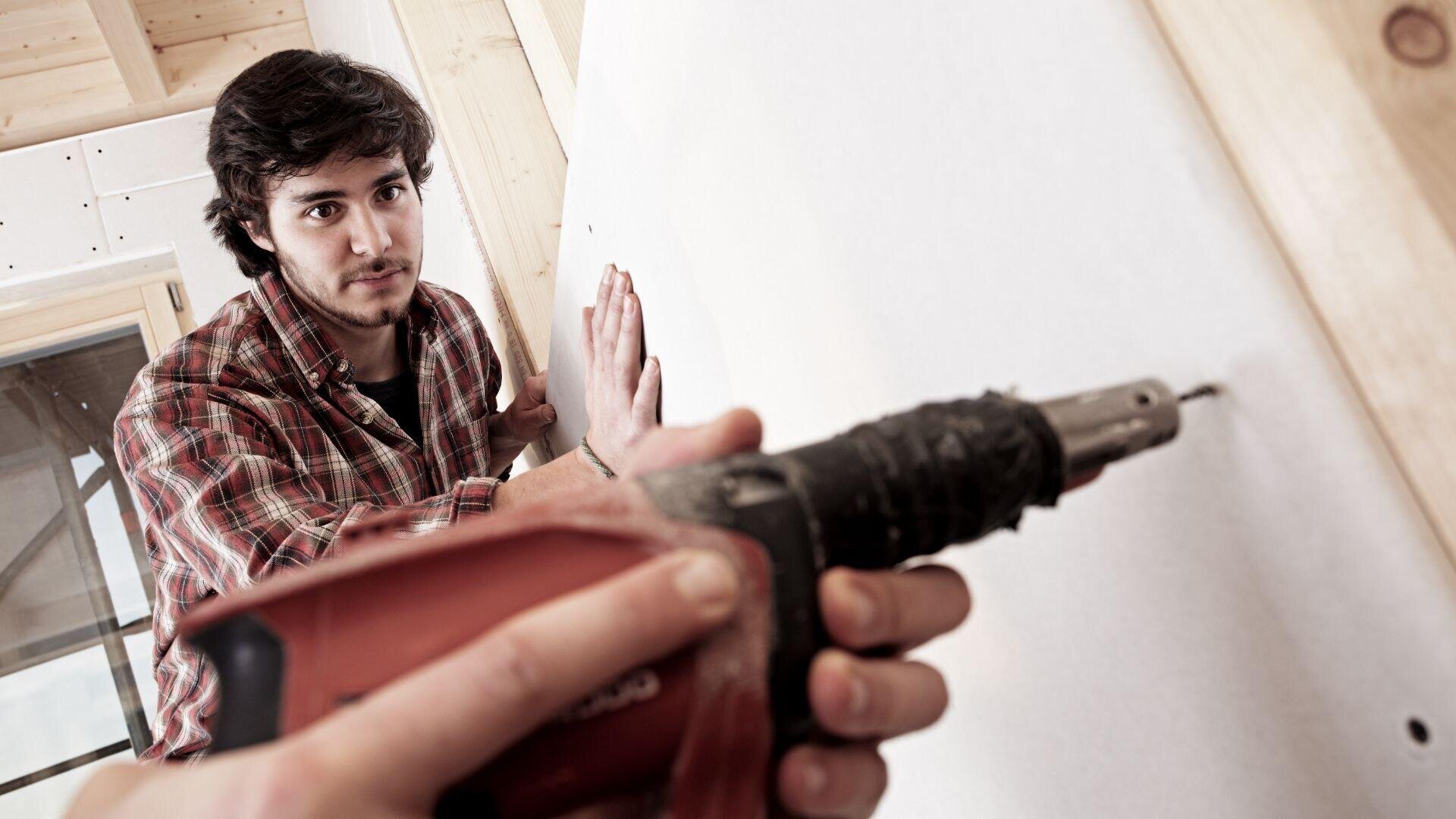 Eine Person im karierten Hemd hält eine Gipskartonplatte fest, während eine andere Person sie bei einer Bau- oder Renovierungstätigkeit mit einer Bohrmaschine an einem Holzrahmen befestigt.