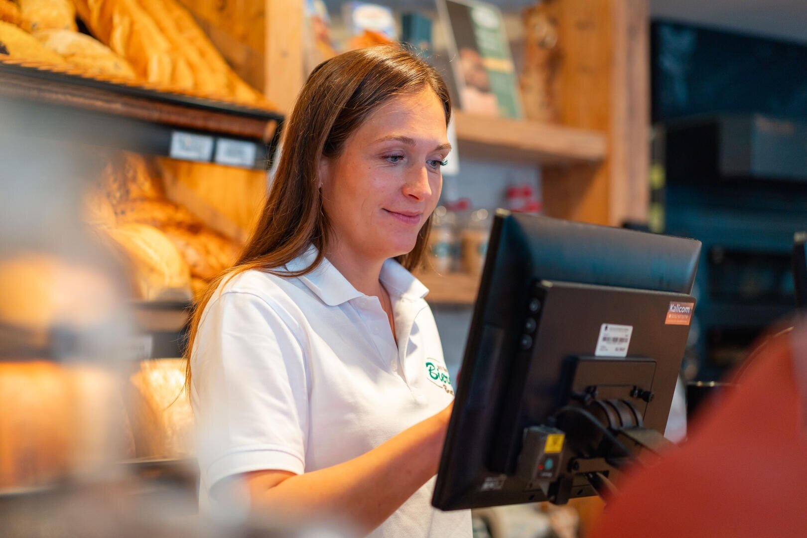Eine Frau in einem weißen Polohemd steht hinter einem Tresen in einer Bäckerei und bedient eine Touchscreen-Kasse. Im Hintergrund sind Regale mit Broten zu sehen.