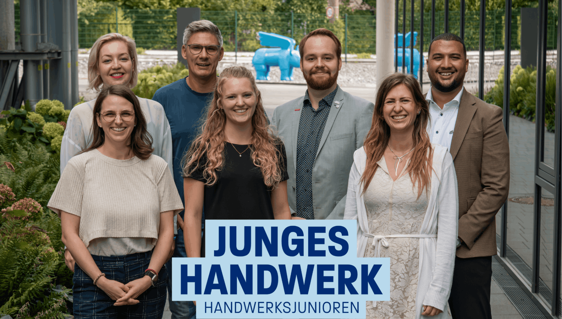 Sieben lächelnde Erwachsene, vier Frauen und drei Männer, stehen gemeinsam draußen vor einer Grünanlage und einem Gebäude. Im Hintergrund ist eine blaue Skulptur zu sehen. Der Text lautet Junges Handwerk Handwerksjunioren.