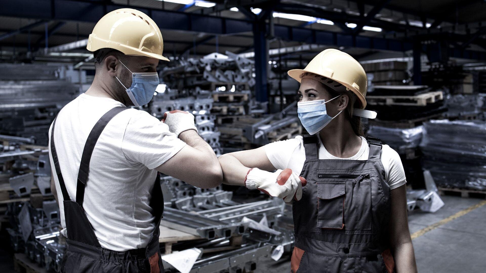 Zwei Fabrikarbeiter mit gelben Schutzhelmen, Gesichtsmasken und Handschuhen begrüßen sich mit einem Ellenbogenstoß in einem Industrielager voller Metallteile und -geräte.