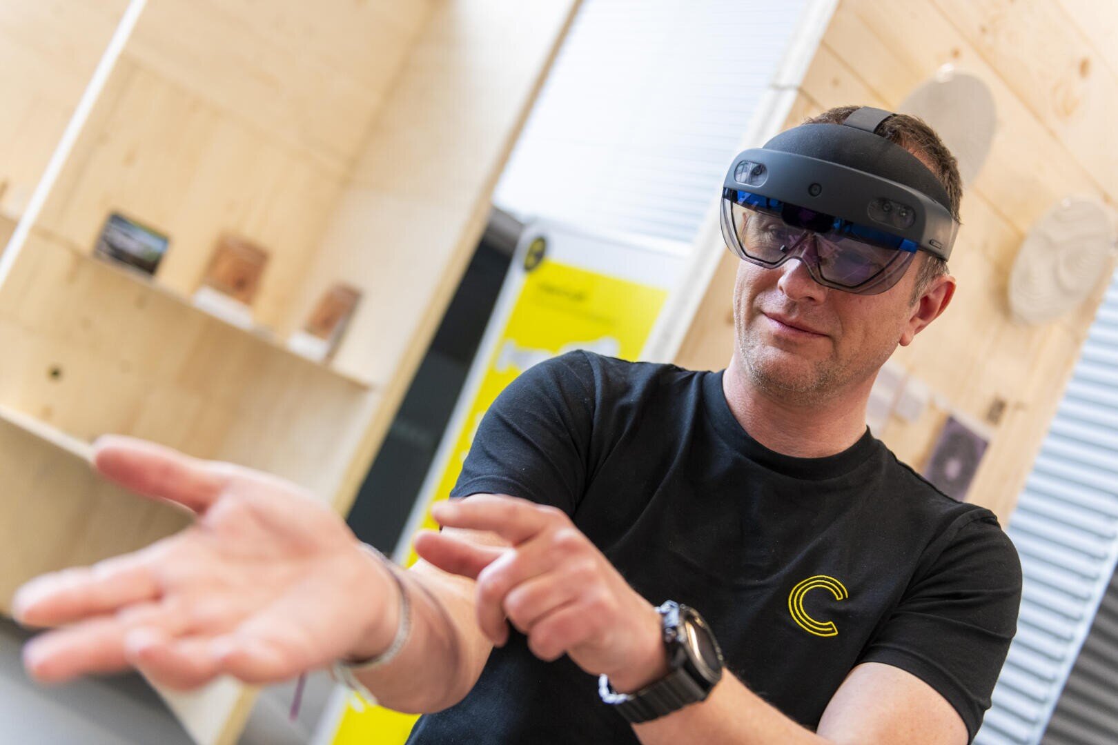 Eine Person, die eine Augmented-Reality-Brille trägt, gestikuliert mit ihren Händen und scheint so mit einem virtuellen Objekt zu interagieren. Sie befindet sich in einem Haus und trägt ein schwarzes Hemd mit einem gelben Logo und eine Smartwatch.