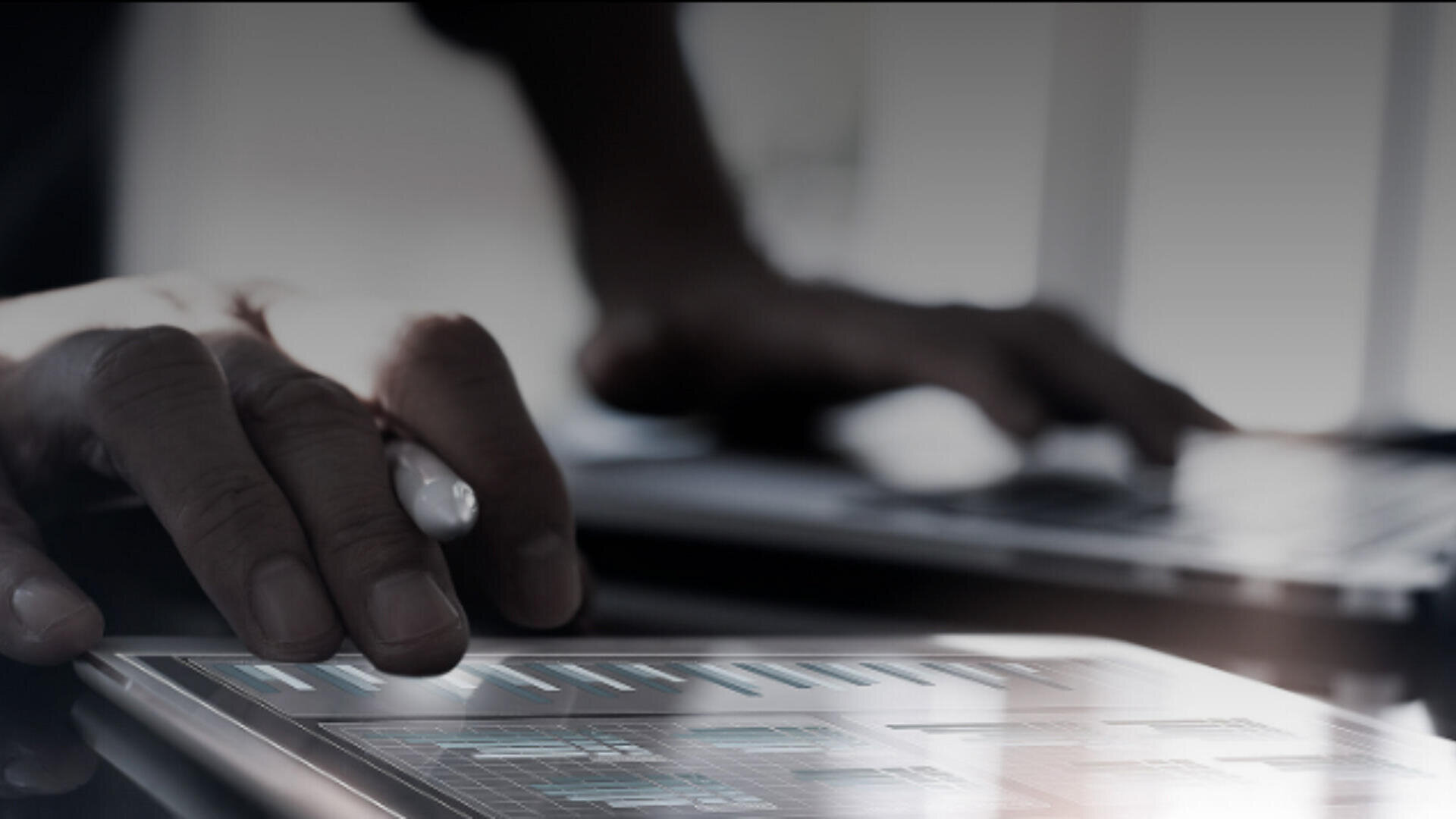 Eine Person benutzt einen Stift auf einem Tablet, auf dem Balkendiagramme angezeigt werden. Im Hintergrund tippt eine andere Hand auf einem Laptop. Das Bild vermittelt eine moderne, technologieorientierte Atmosphäre.