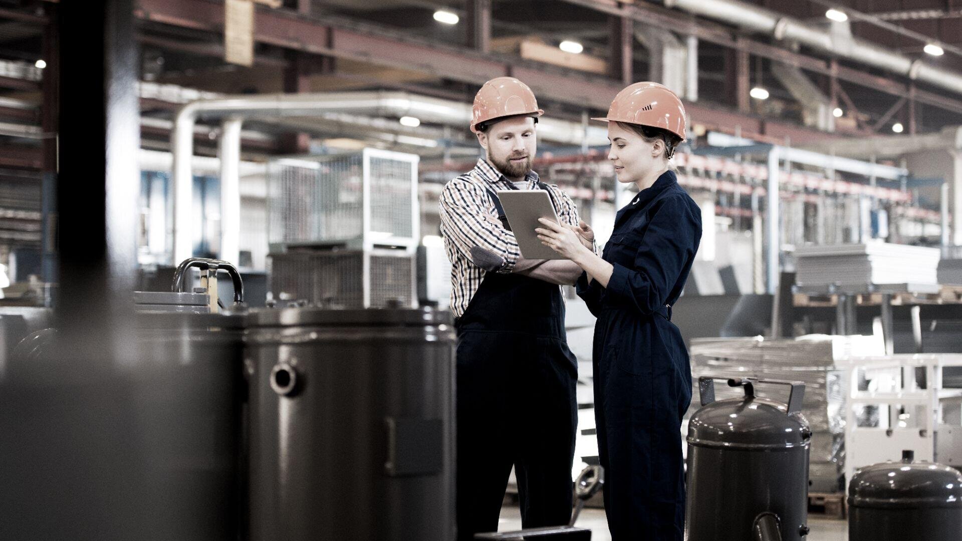 Zwei Fabrikarbeiter mit orangefarbenen Schutzhelmen und Uniformen stehen in einer Industrieumgebung und besprechen etwas auf einem Klemmbrett, umgeben von großen Maschinen und Metallgeräten.