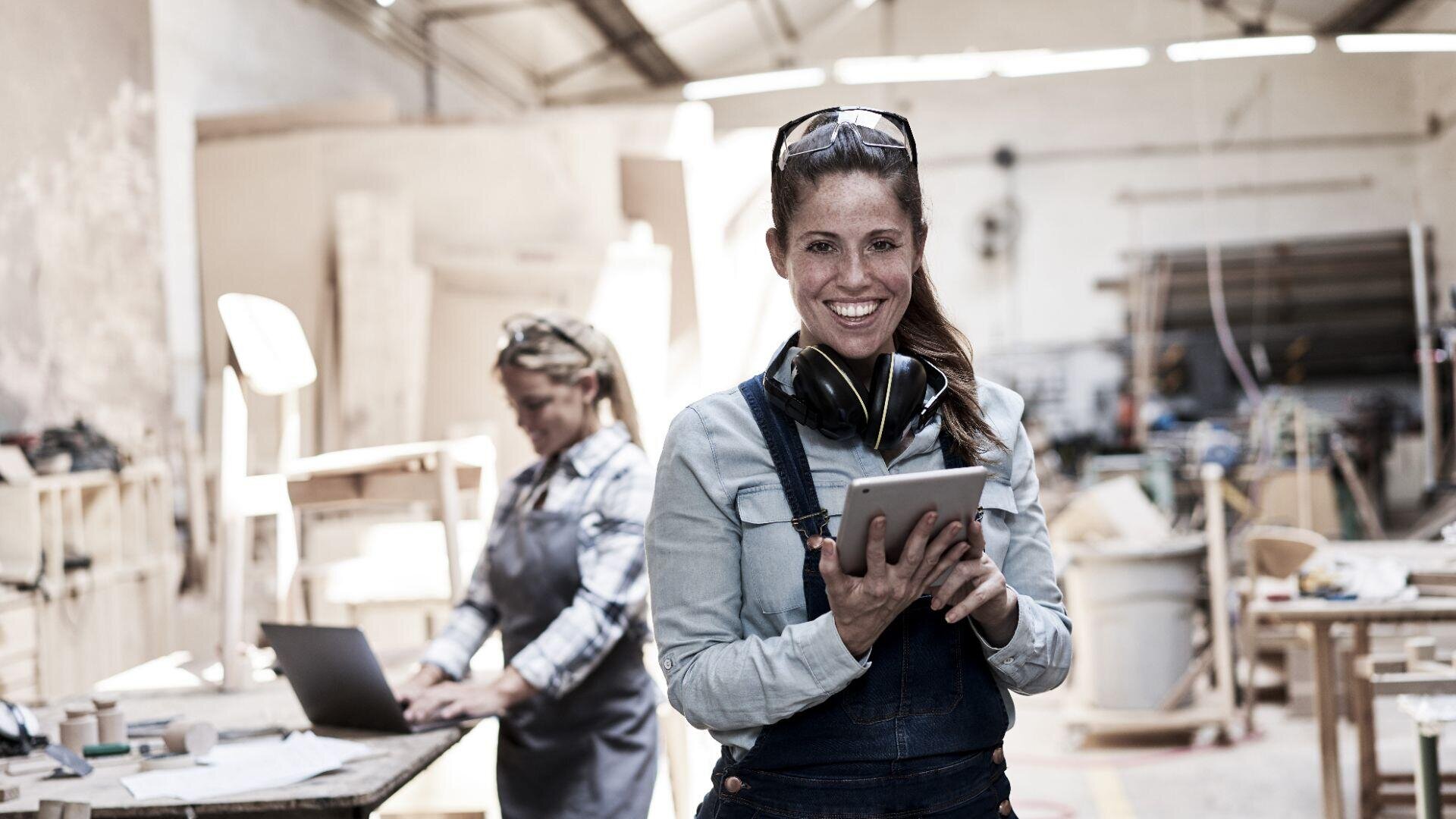 Eine Frau in Overall und Schutzausrüstung lächelt in einer Holzwerkstatt und hält ein Tablet in der Hand. Im Hintergrund arbeitet eine andere Person an einem Laptop, umgeben von Werkzeugen und Holzmaterialien.