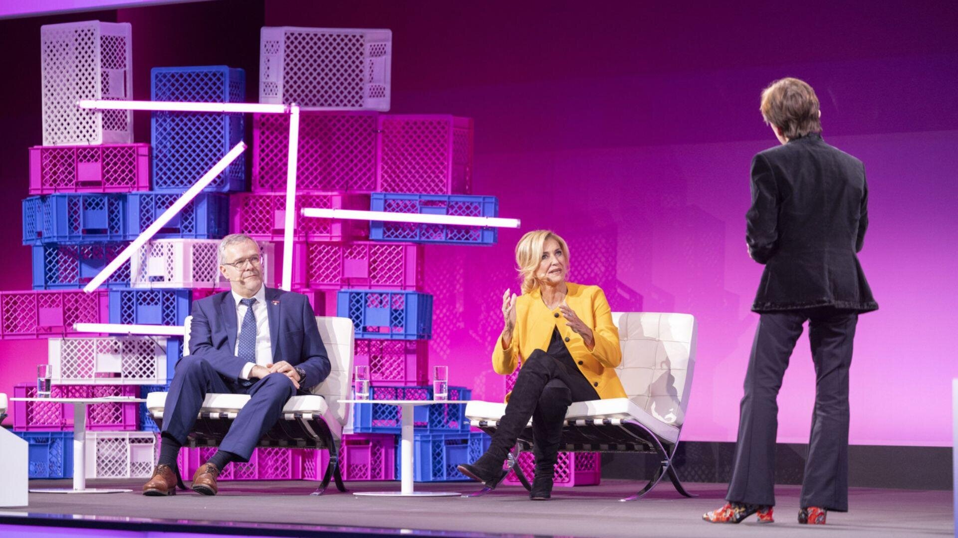 Drei Personen stehen auf einer hell erleuchteten Bühne mit weißen Stühlen und lila-rosa kistenartigen Strukturen im Hintergrund. Zwei sitzen, einer trägt einen blauen Anzug, einer eine gelbe Jacke, während ein dritter ihnen gegenübersteht.