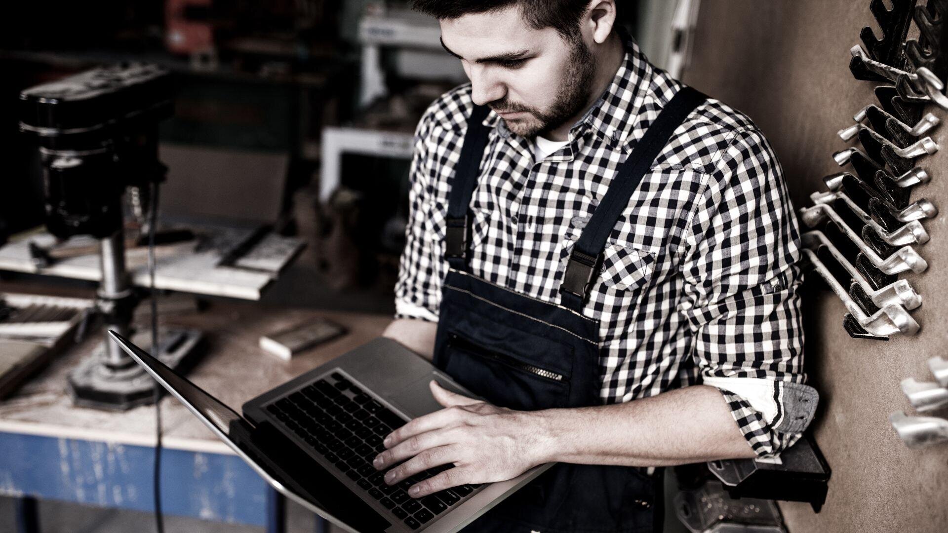 Ein Mann in kariertem Hemd und Overall steht in einer Werkstatt und bedient einen Laptop. Im Hintergrund sind verschiedene Werkzeuge und Geräte zu sehen, die auf eine Tischlerei oder Holzverarbeitung hindeuten.