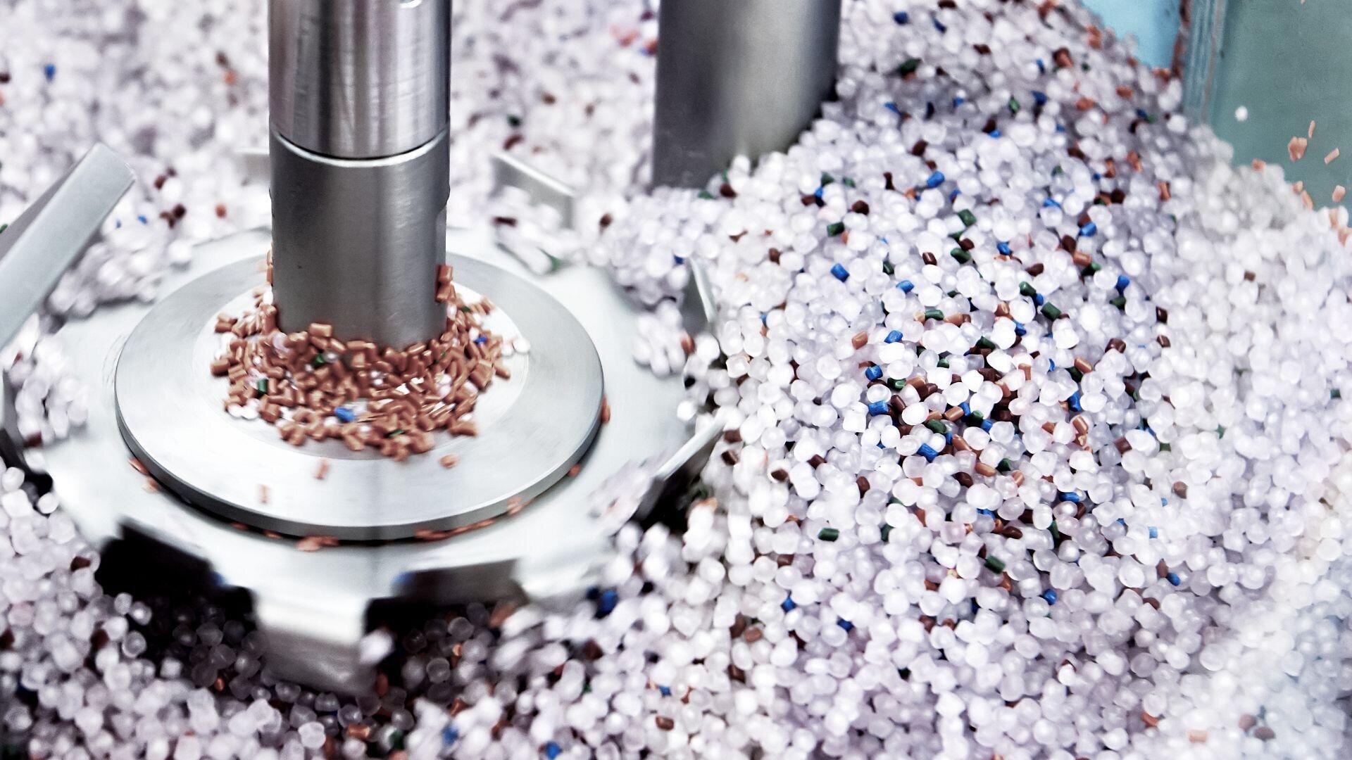 Nahaufnahme einer Maschine, die in einer industriellen Umgebung eine Mischung aus kleinen, runden Kunststoffpellets in verschiedenen Farben, darunter Weiß, Braun und Blau, rührt.