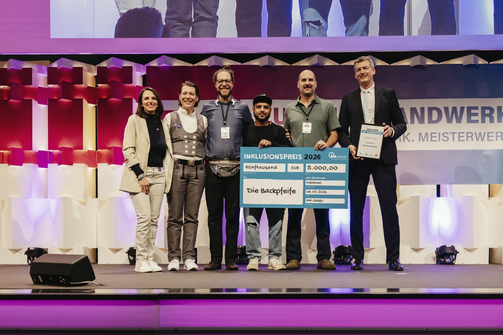Sechs Personen stehen lächelnd auf der Bühne, in der Hand einen großen Scheck über 5.000 Euro mit der Aufschrift Inklusionspreis 2026 und Die Backpfeife. Ganz rechts steht ein Mann im Anzug. Im Hintergrund sind bunte Lichter und Bühnendekorationen zu sehen.