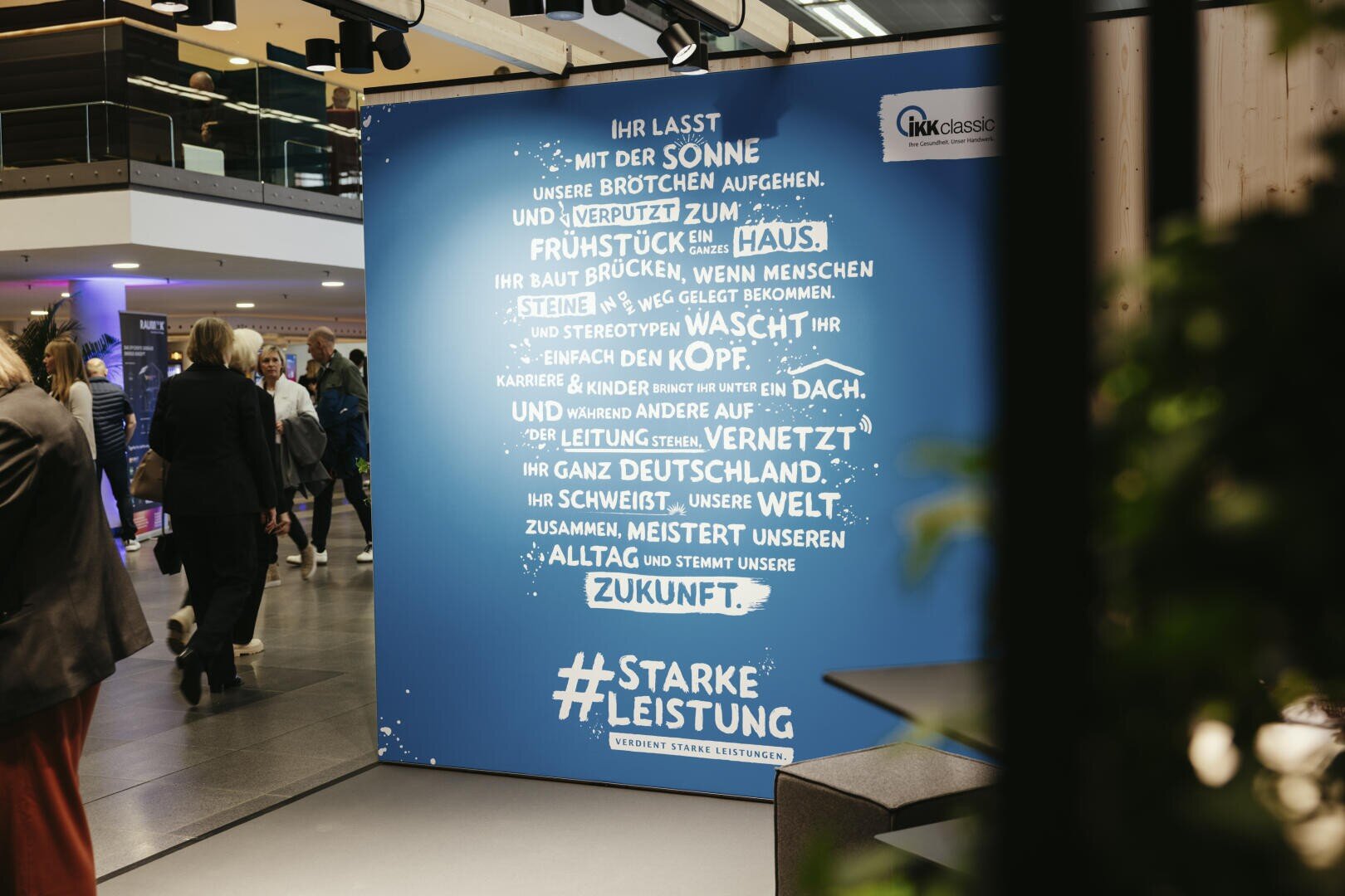 Auf einer blauen Anzeigetafel in einem öffentlichen Innenbereich ist ein deutscher Text in Form eines Hauses zu sehen, der die Themen Stärke und Gemeinschaft hervorhebt. Im Hintergrund gehen und stehen mehrere Menschen.