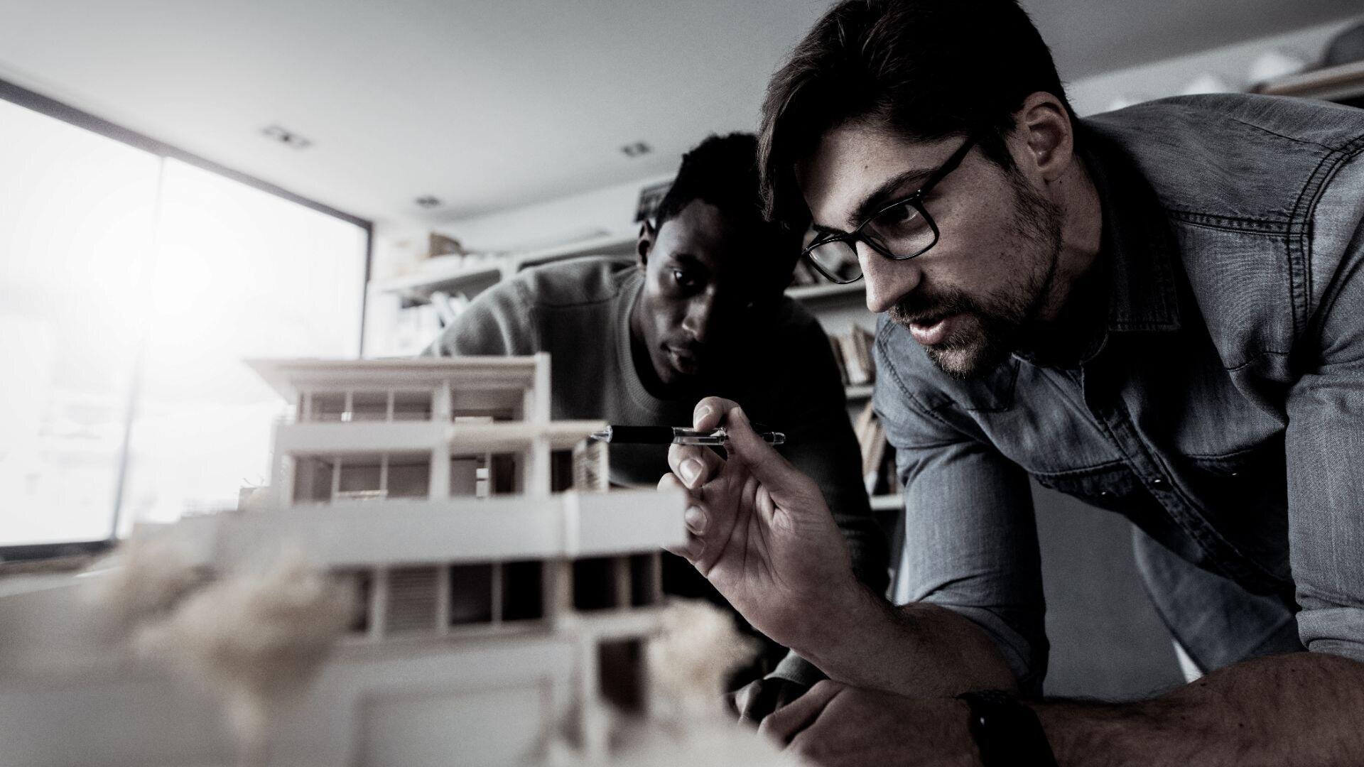 Zwei Männer untersuchen und arbeiten im Innenbereich genau an einem detaillierten Architekturmodell eines modernen Gebäudes. Einer hält ein Werkzeug in der Hand und trägt eine Brille. Beide wirken konzentriert und vertieft in ihre Aufgabe.