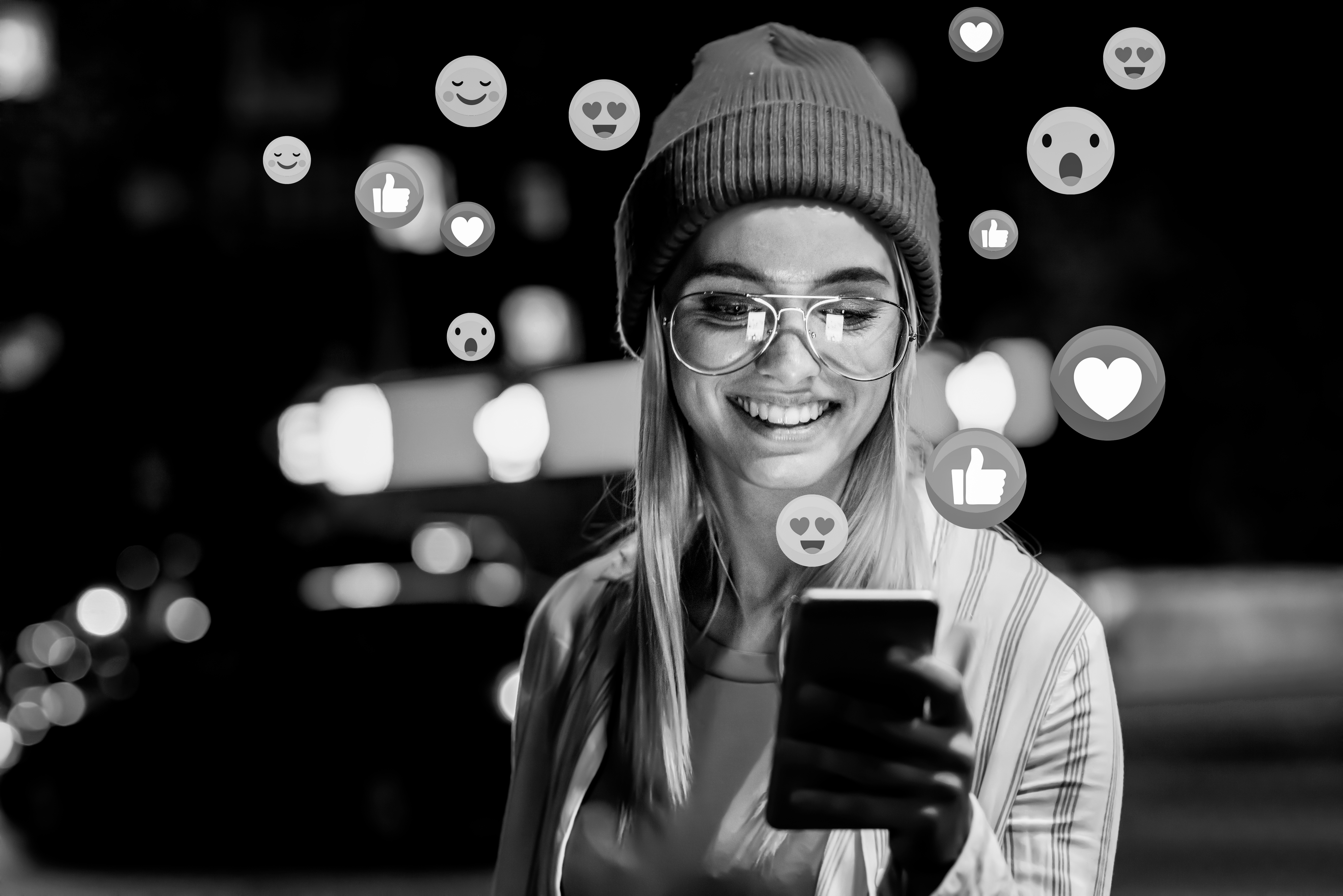Eine junge Frau mit Brille und Mütze lächelt nachts auf ihr Telefon. Symbole für Reaktionen in sozialen Medien wie Herzen, Daumen hoch und Smileys werden um sie herum eingeblendet.